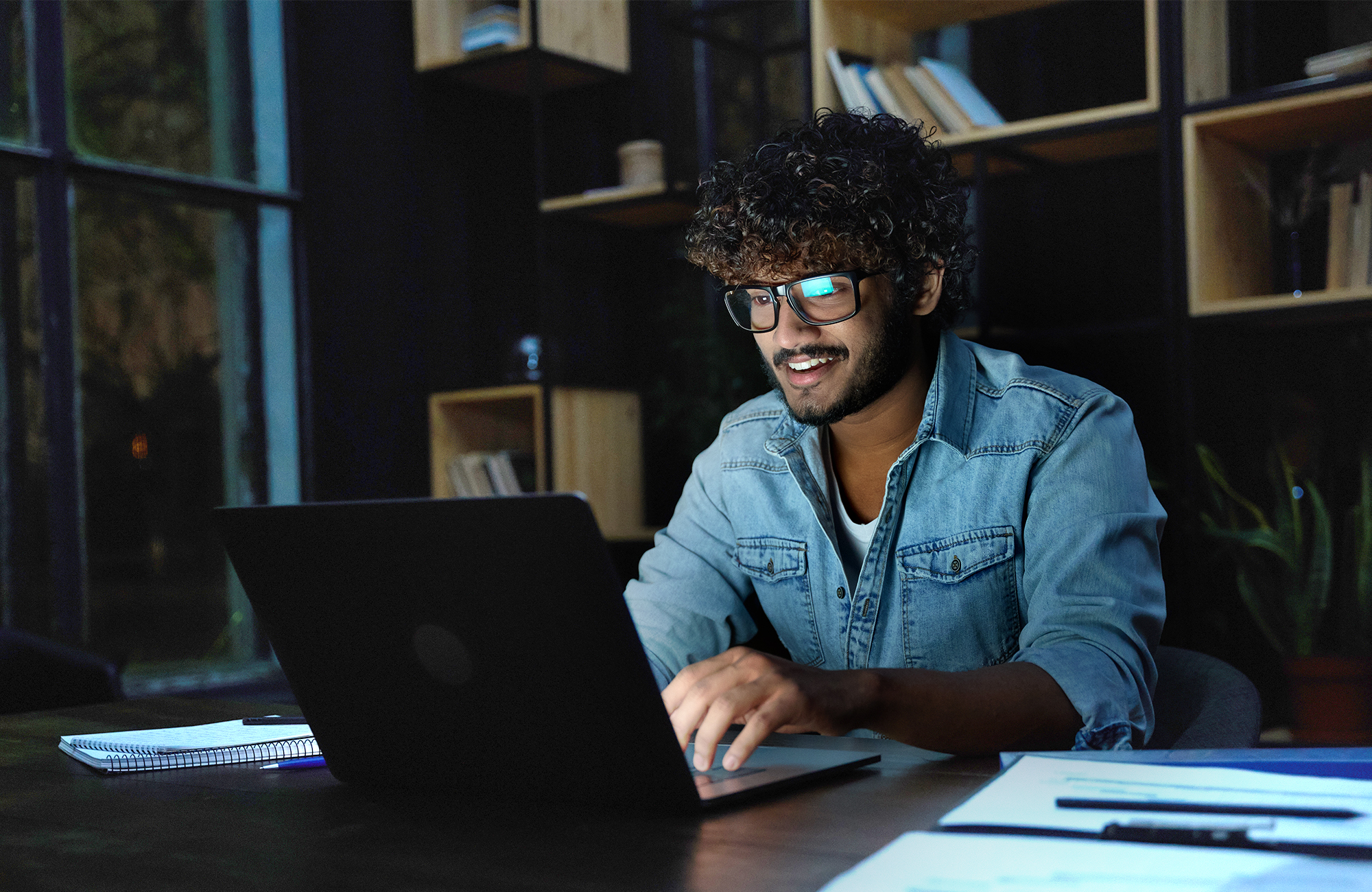 Person in a denim shirt working on a laptop at a desk, bookshelves and window in the background.